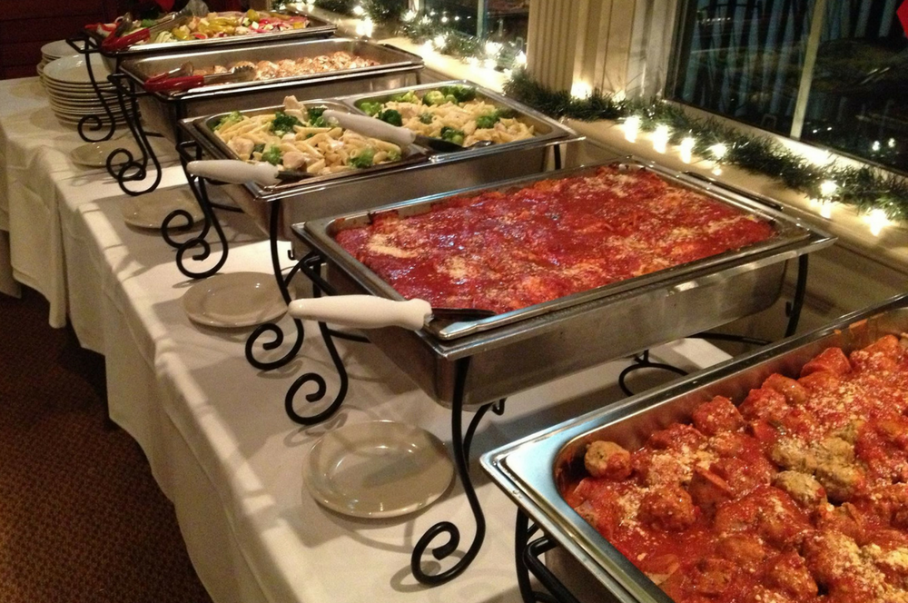 Buffet table with various dishes: pasta, meatballs, sauce. White tablecloth, plates, holiday lights in the background.