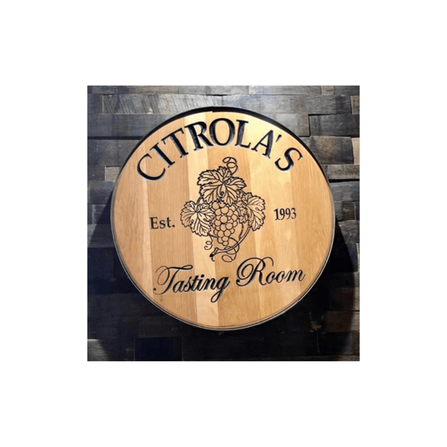 Citrola's Tasting Room sign, round wood with etched grape design, mounted on a brick wall.