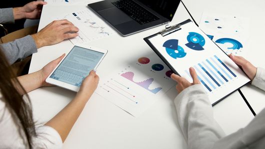 People at a table review data on a laptop, tablet, and charts with blue and white graphs.