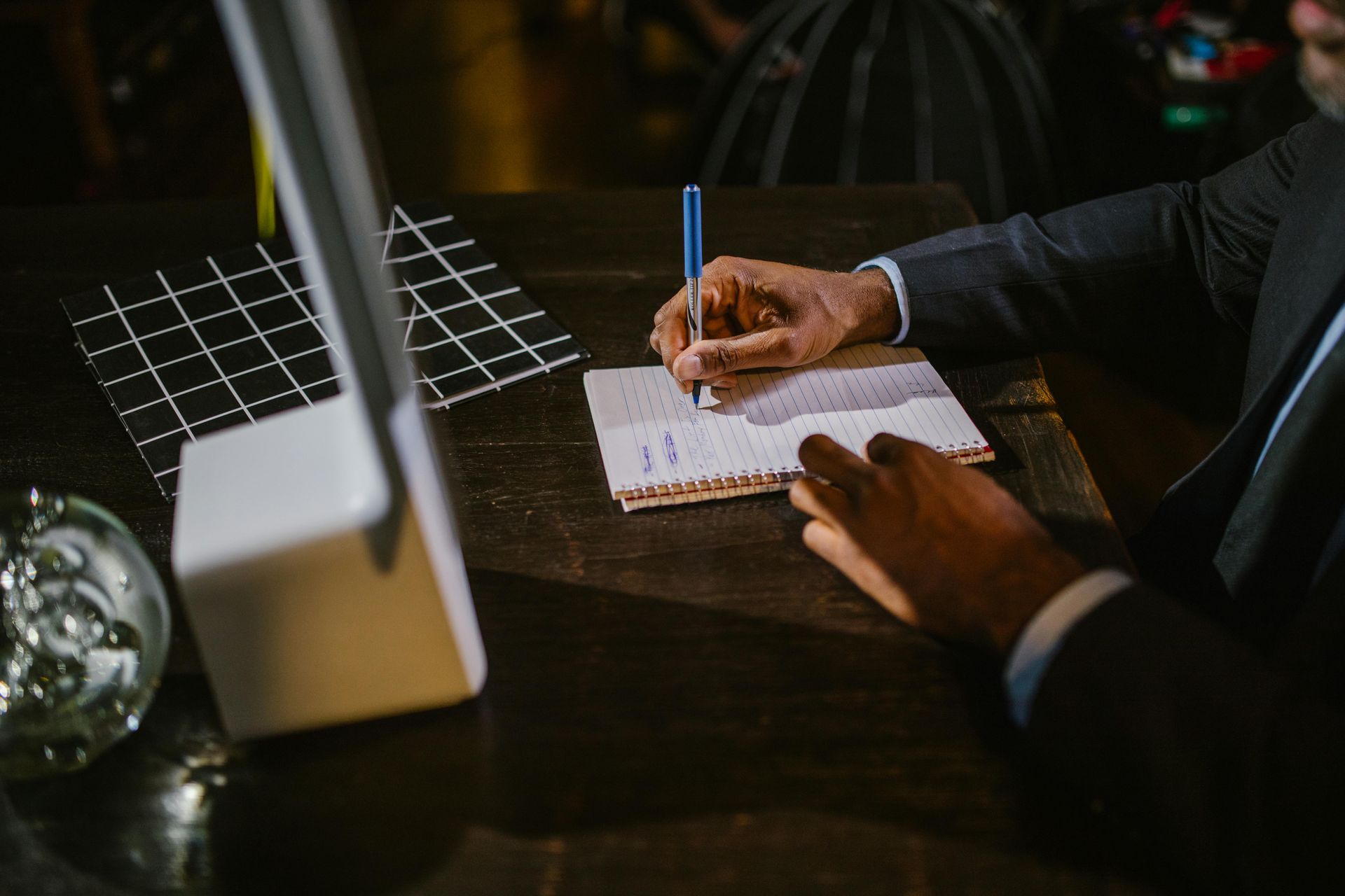 Person in suit writing on a notepad at a desk, with a lamp and geometric decor.