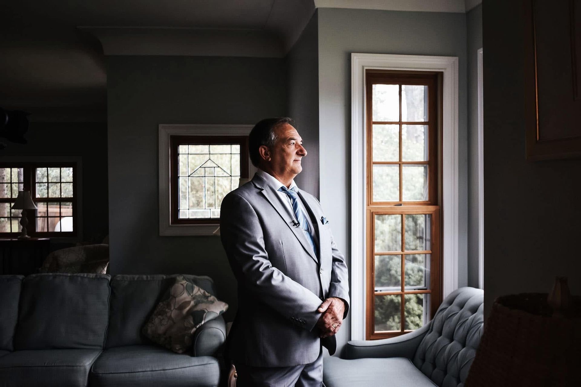 A man in a suit and tie is standing in front of a window in a living room.