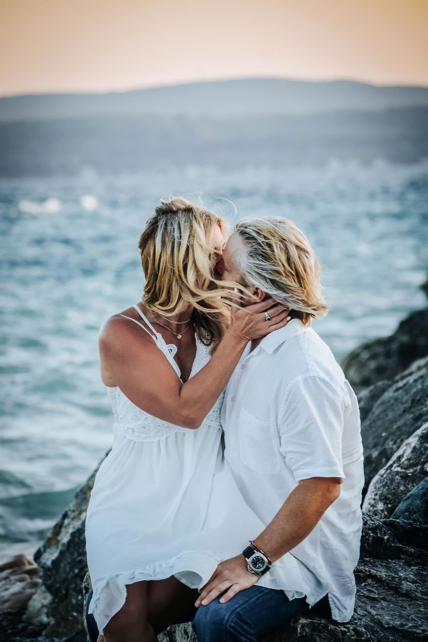 A man and a woman are kissing on a rock near the ocean.