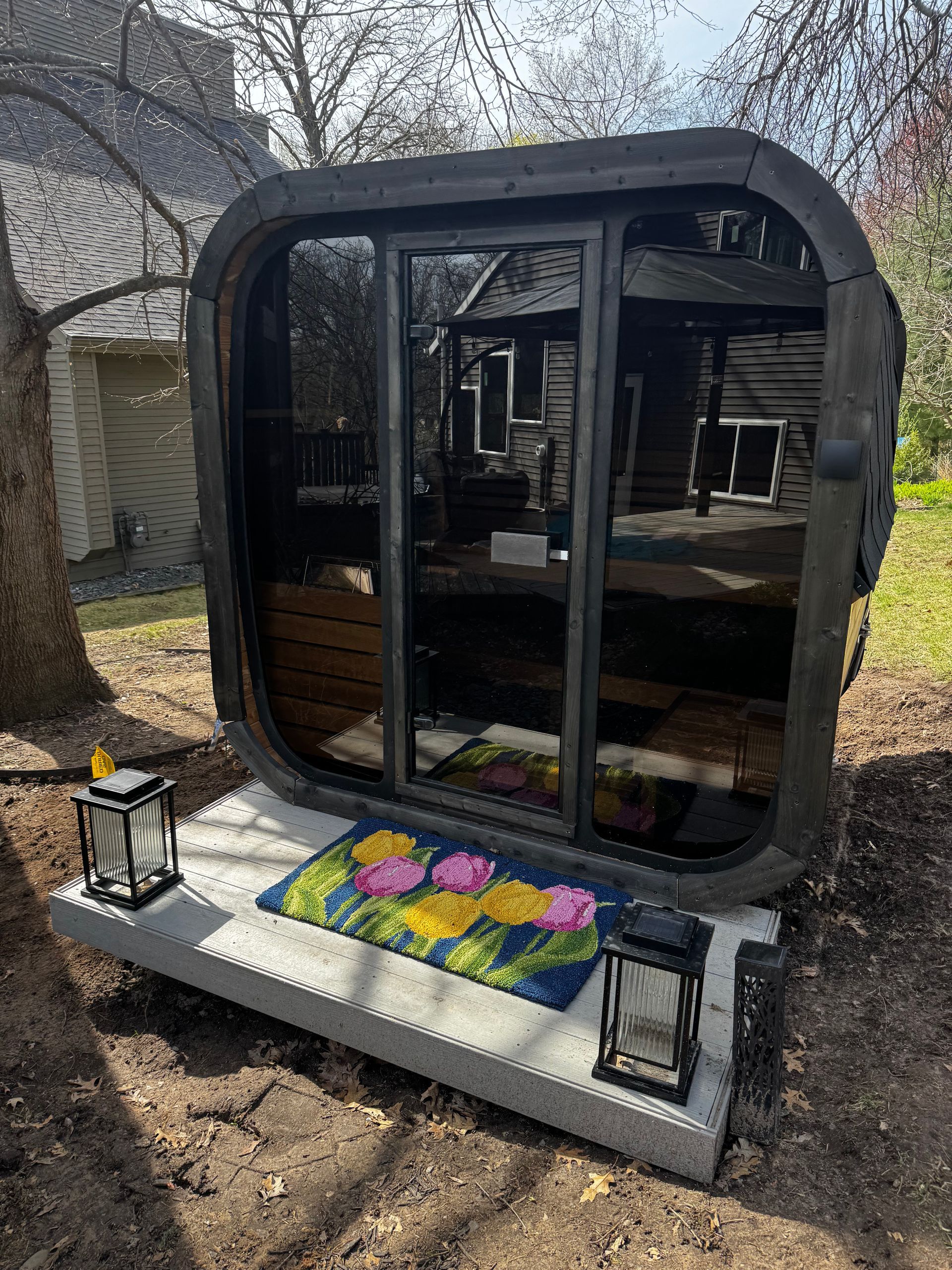 Small black micro-cabin with glass doors, standing on a concrete pad with flower decorations in a wooded yard