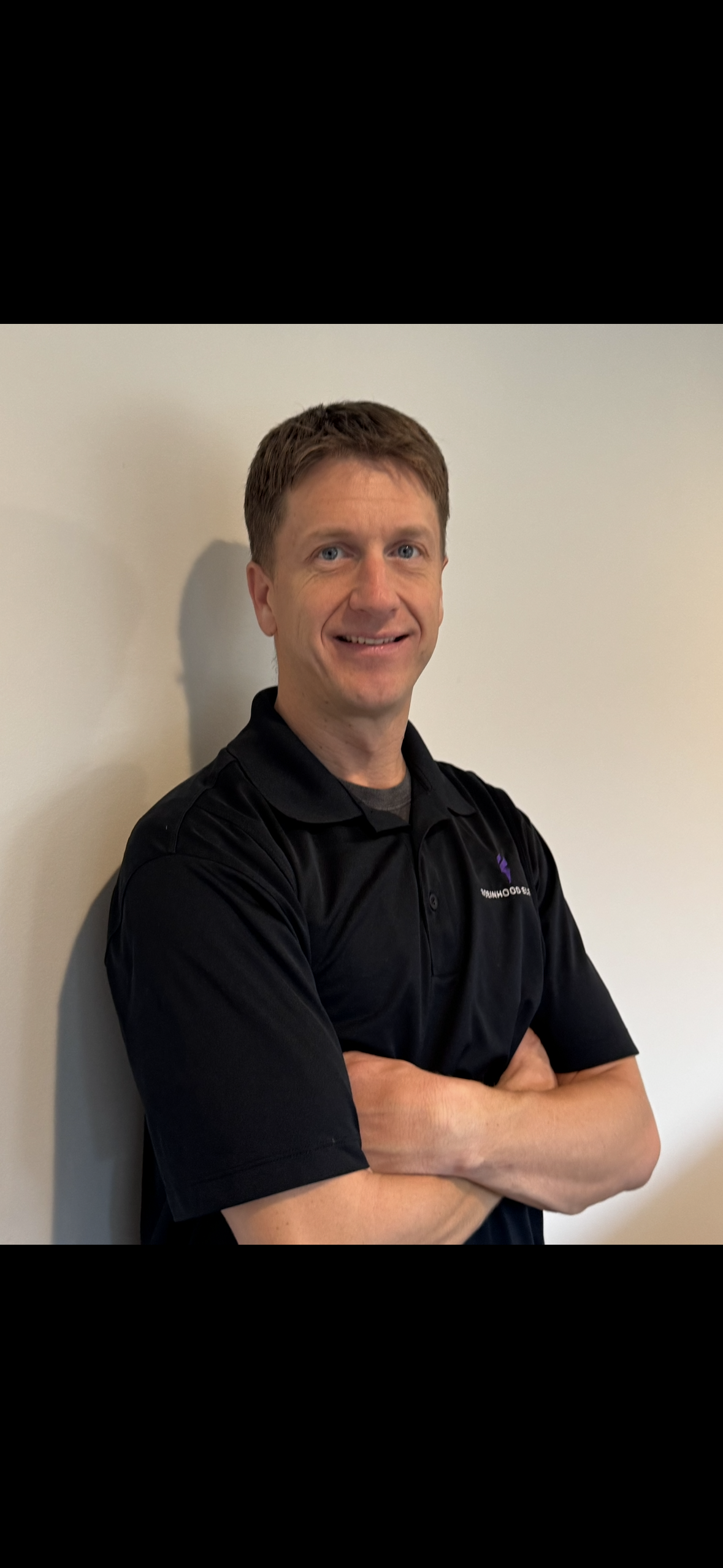 Smiling man in a black polo shirt standing with arms crossed against a plain wall.