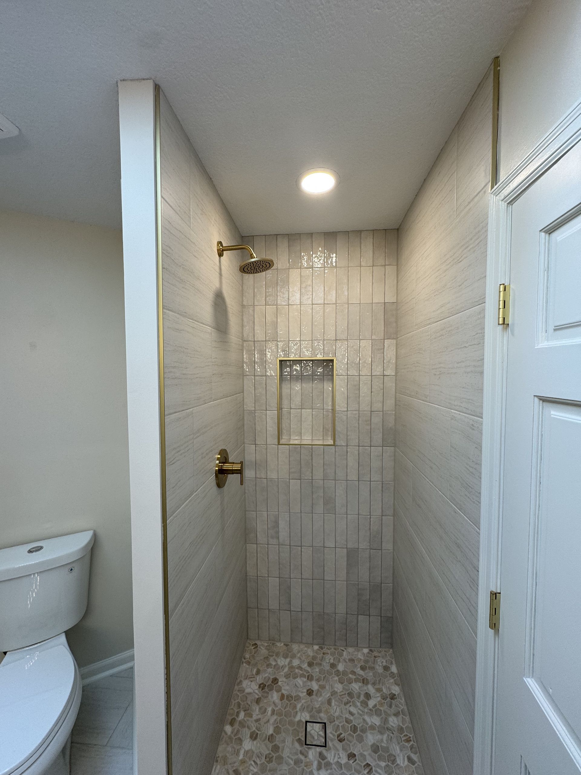 Small beige-tiled bathroom shower with glass door and recessed light, next to a toilet.