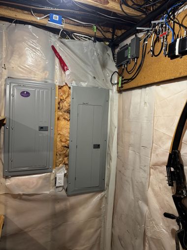 Basement wall with two gray electrical panels, exposed insulation, and hanging wires.