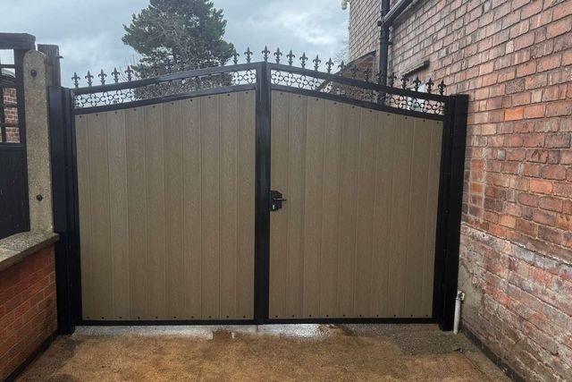 Natural oak coloured composite driveway gates in a black metal frame with fleur-de-lis finials and decorative scrollwork