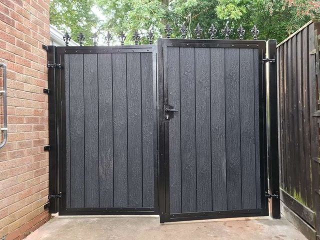 Black composite double gates with fleur de lis finials fitted in a passage between a house and fence
