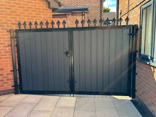 Dark composite driveway gates topped with cast fleur de lis finials between brick pillars