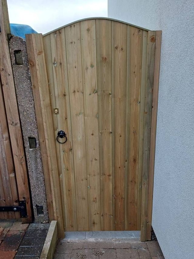 Tall arched top tongue and groove garden gate in a narrow passage between a timber post and a rendered house wall