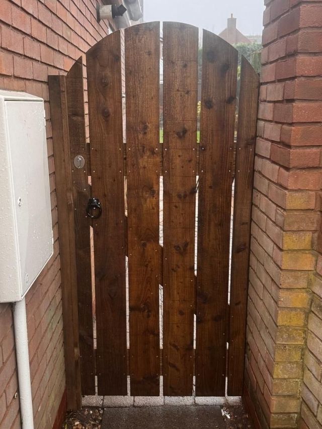Dark stained bespoke arched side gate with spaced vertical boards, hung between a house wall and a brick pier