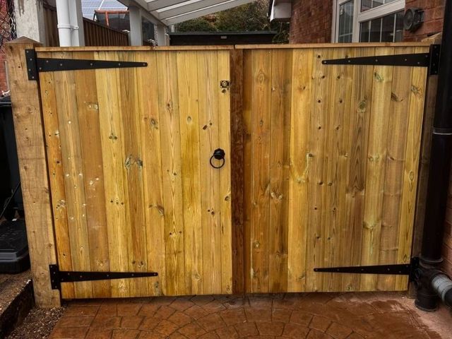 Wooden double gates closing a carport opening, seen from outside with tee hinges and a ring handle on a patterned drive