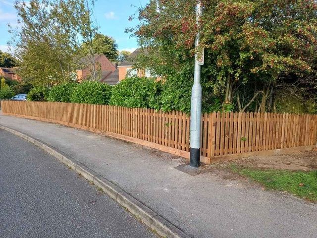 Brown treated picket fencing along a pavement in front of a laurel hedge beside a residential road