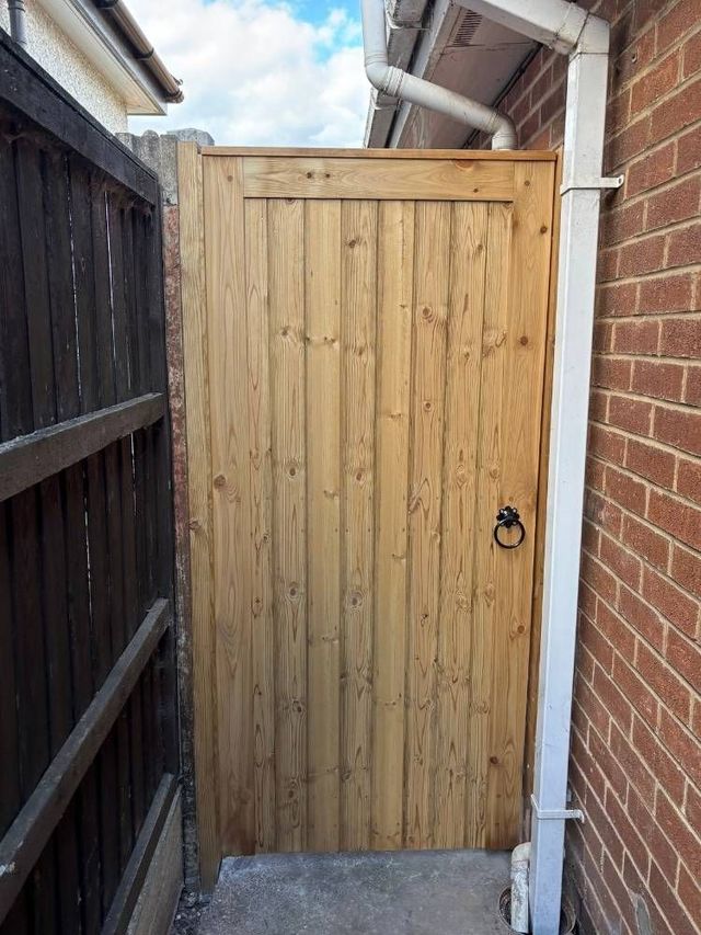 Tongue and groove wooden garden gate hung between an older dark fence panel and a brick house wall with a downpipe