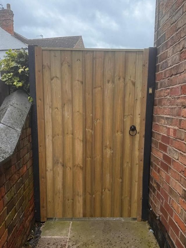 Wooden side garden gate hung on dark painted frame timbers fixed between two brick walls over a flagged path