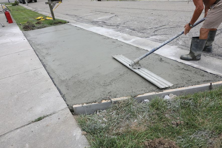 Worker smoothing fresh concrete sidewalk at a street corner with a long-handled screed