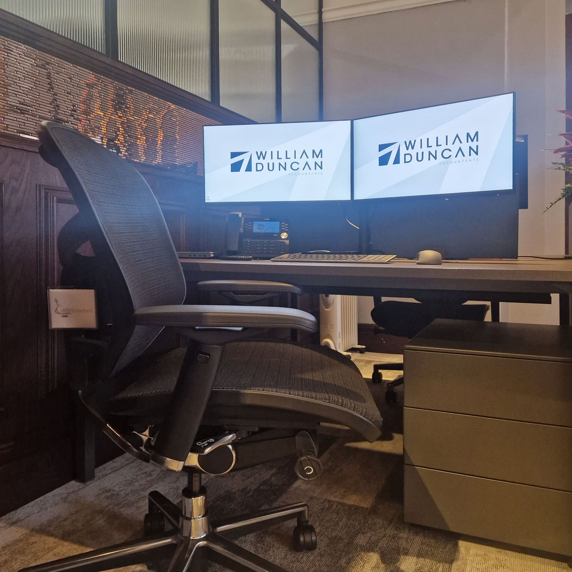 A black ergonomic chair sits at a desk with two monitors on it, showcasing the William Duncan logo.