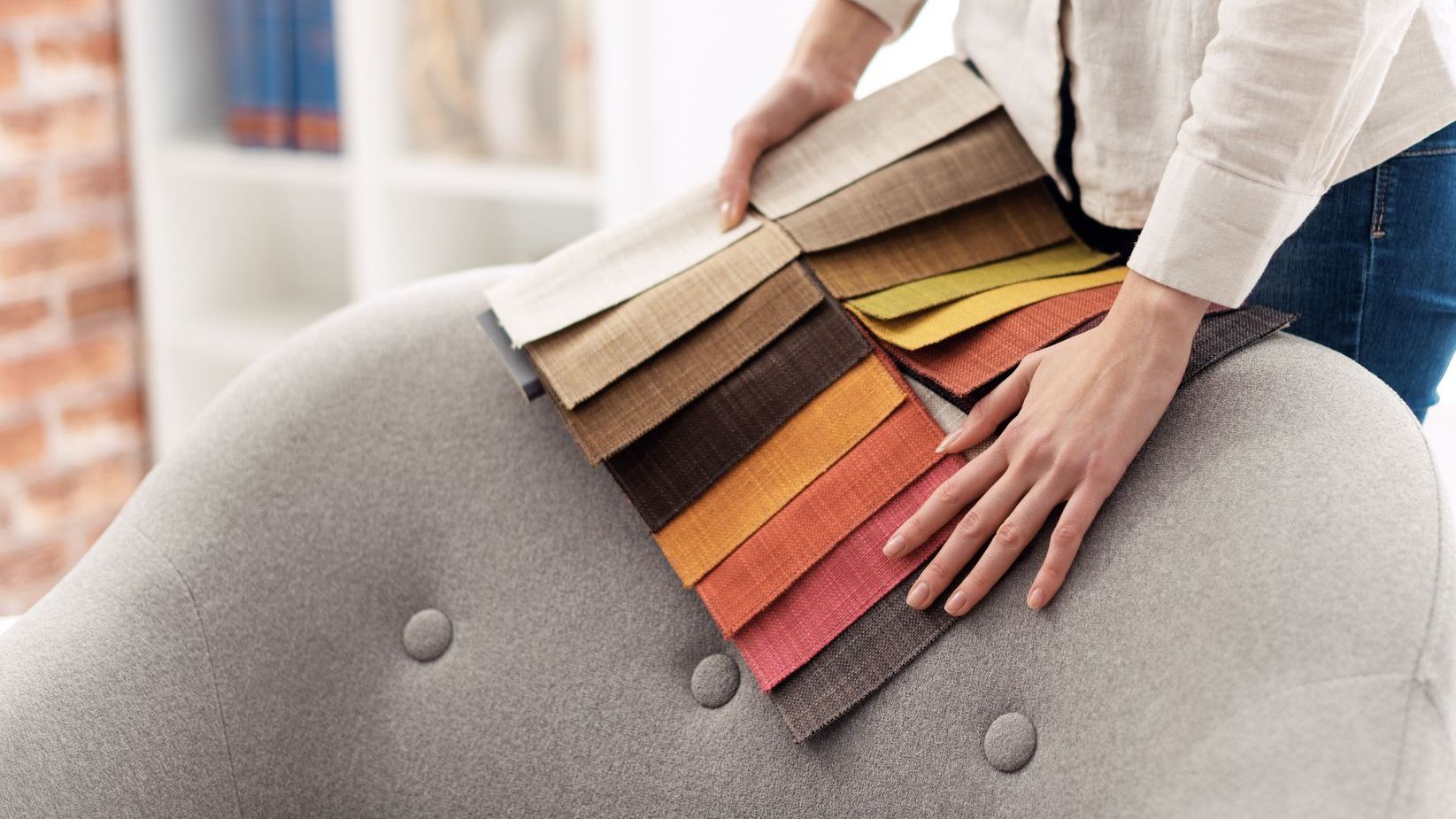 Person holding fabric swatches over a gray upholstered chair, selecting colors for upholstery.