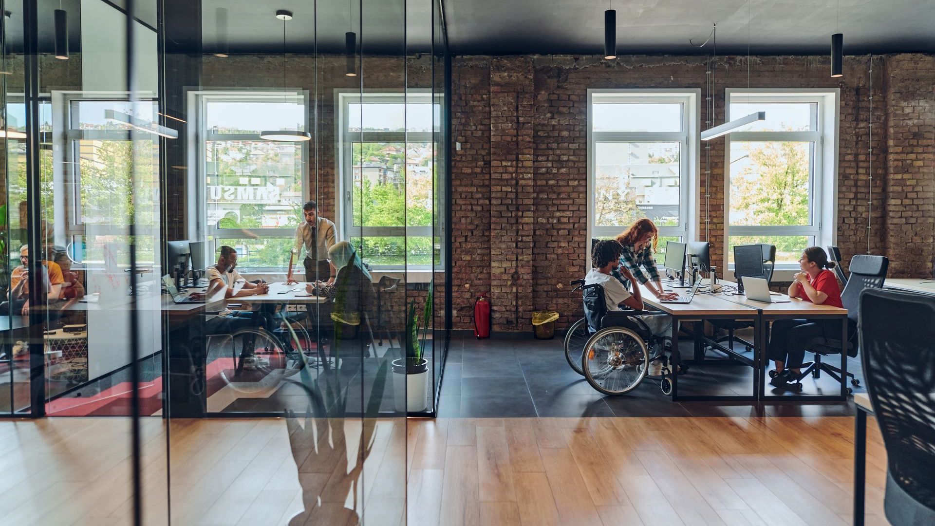 A modern office space with wooden flooring, tile flooring and brick walls. There are bench desks that suit wheelchair users and office chairs. Large open windows let in natural lighting.