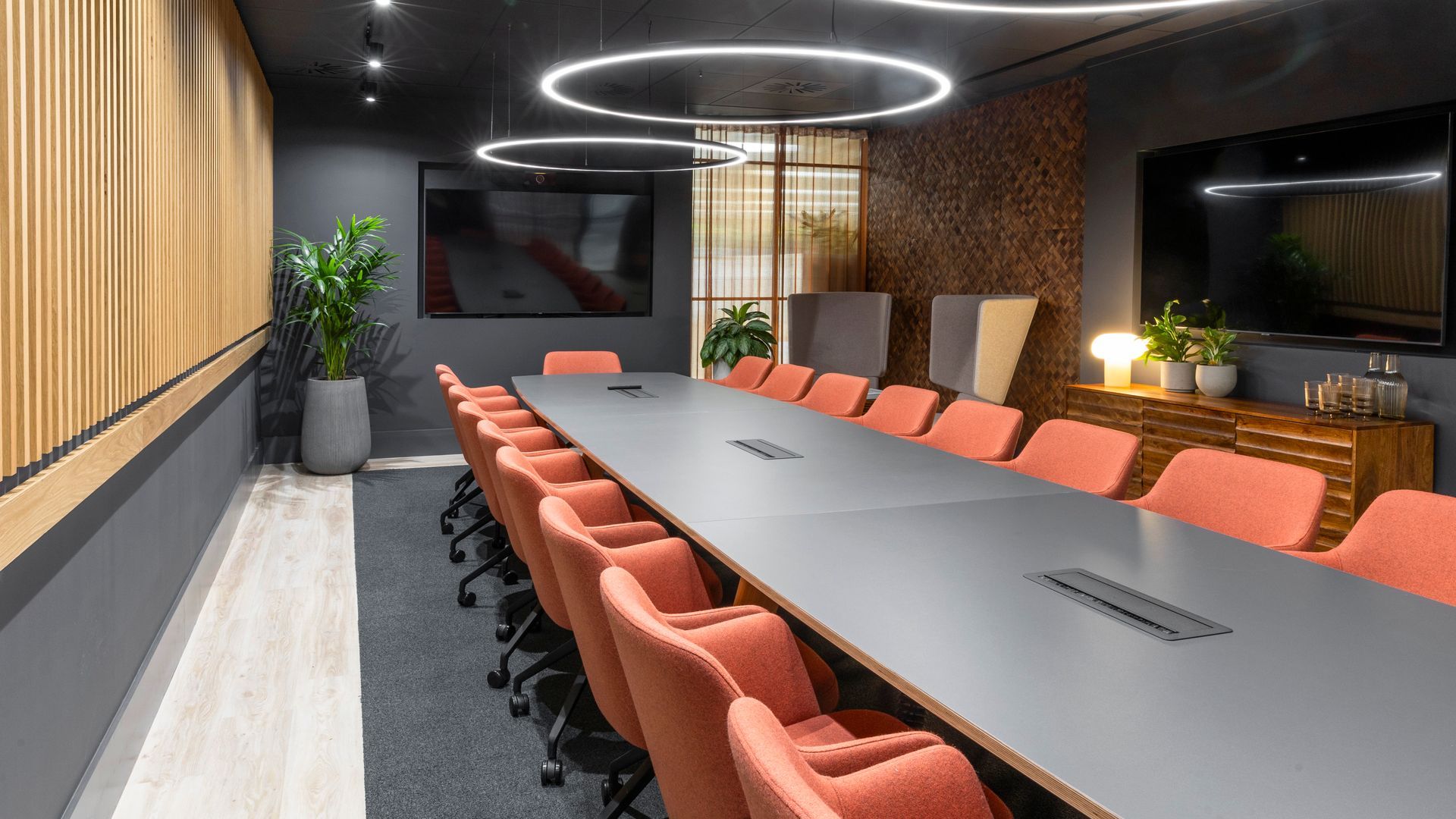 Conference room with long table, red chairs, and two wall-mounted TVs.
