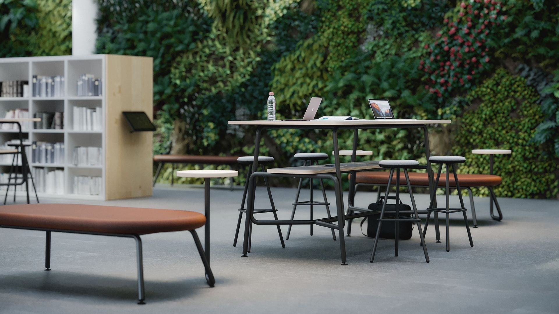 there is a black and white table with stools in the centre of the image. on the table is a water bottle, and two laptops in the background there is a free-standing bookcase full of black, blue and white books. the wall behind the scene is covered in thick and lush greenery. 