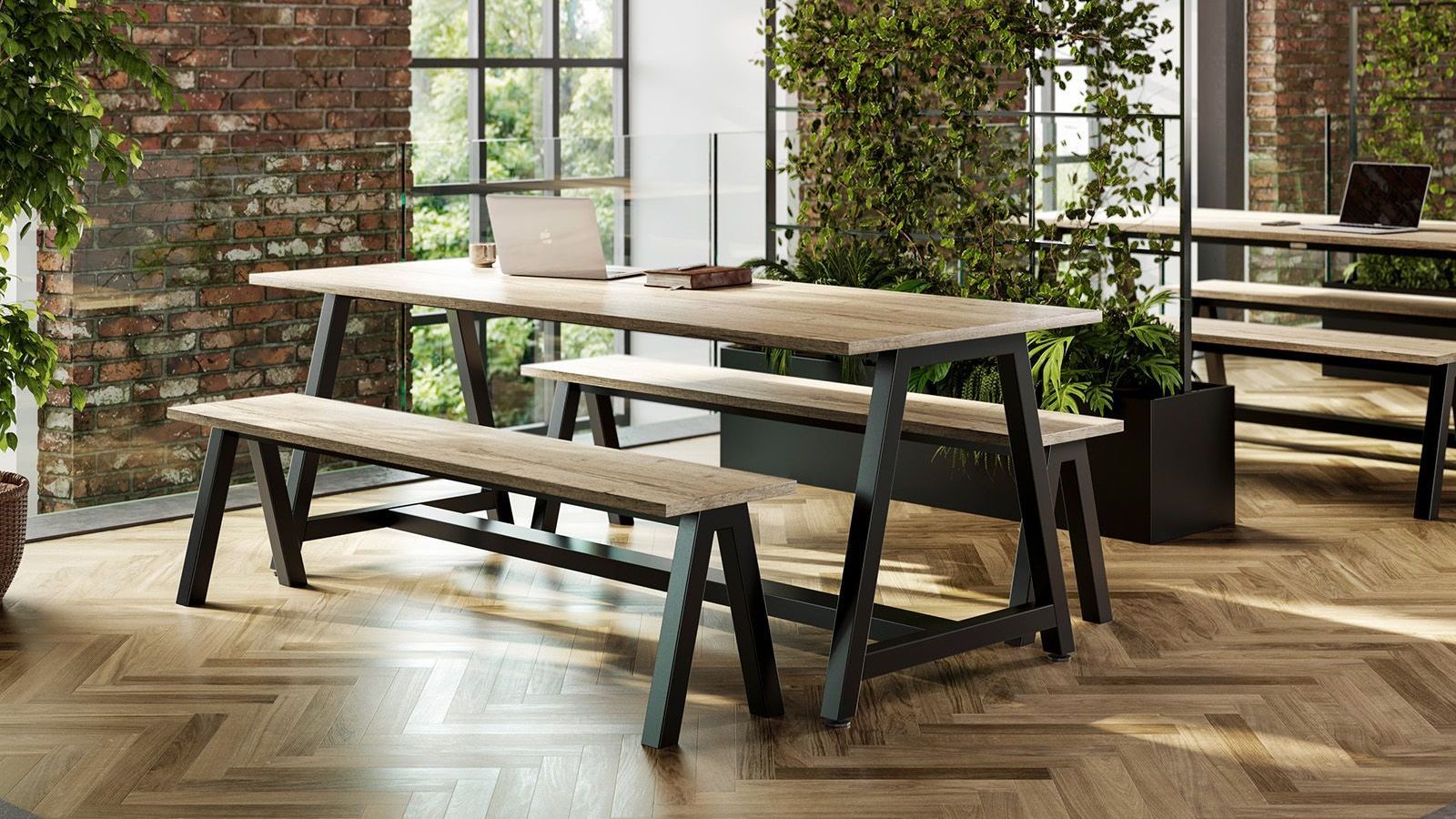 wood picnic table with black legs is at the centre of the photo. the table is indoors in a naturally lit office with brick walls, herringbone flooring, and living dividers. on top of the table is an old book, laptop and mug. 
