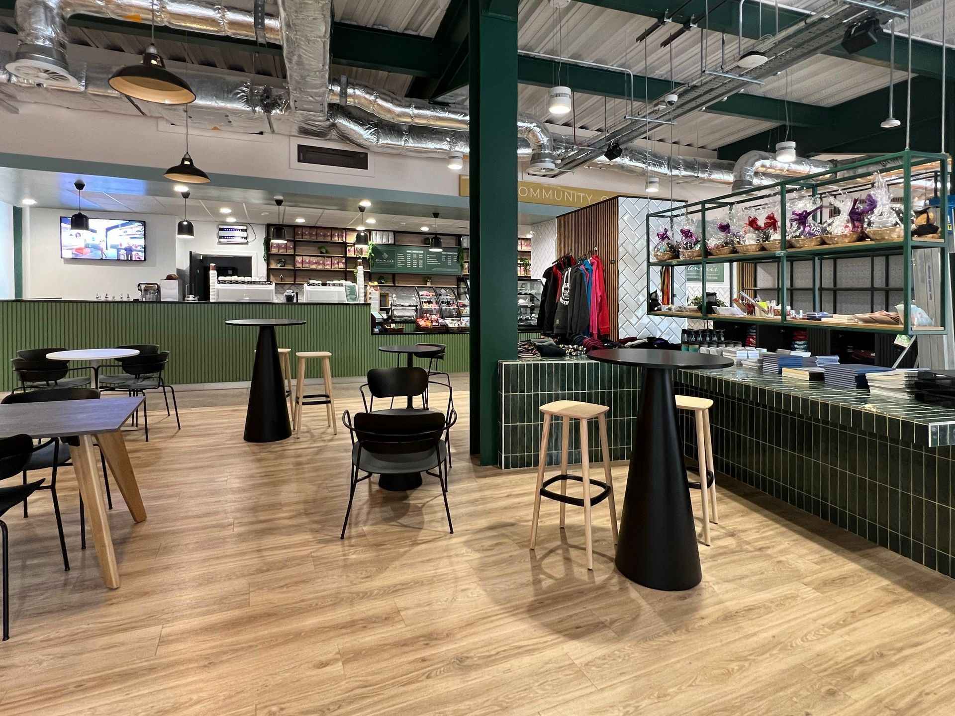 Interior of Annie's Cafe, focusing on the mixture of colours, textures, and furniture choices, including tall black tables, wooden stools, a green tiled retail counter, black dining chairs, and a green panelled serving counter to purchase food.