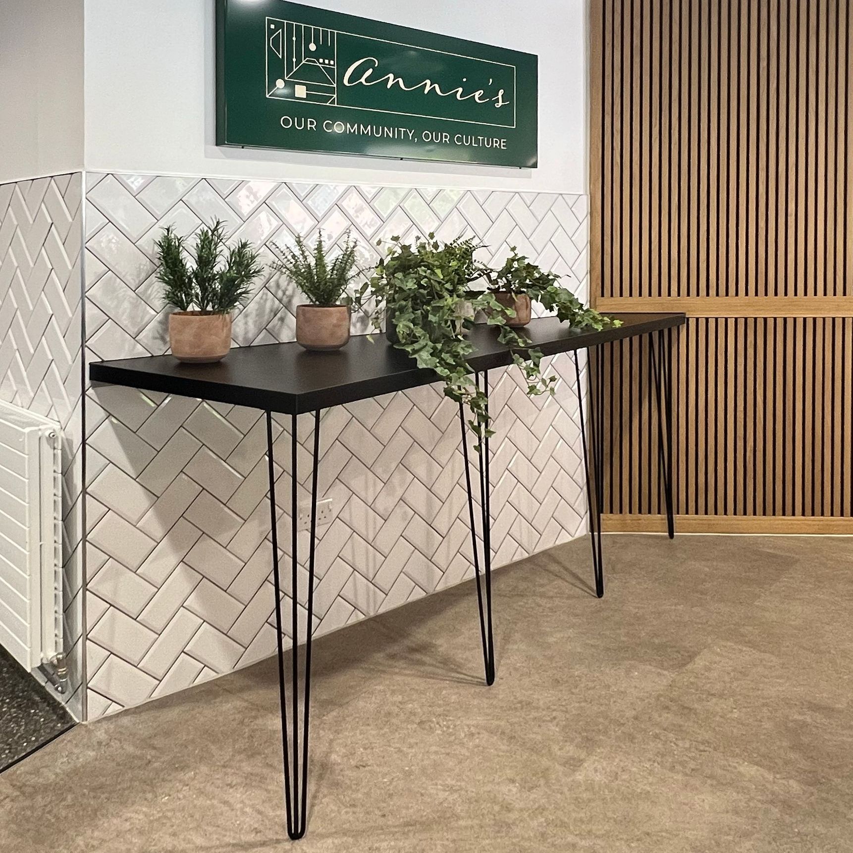 A black table with hairpin legs against a white tiled wall. On the table are four plants. Above the table on the wall is the sign for Annie's Cafe.