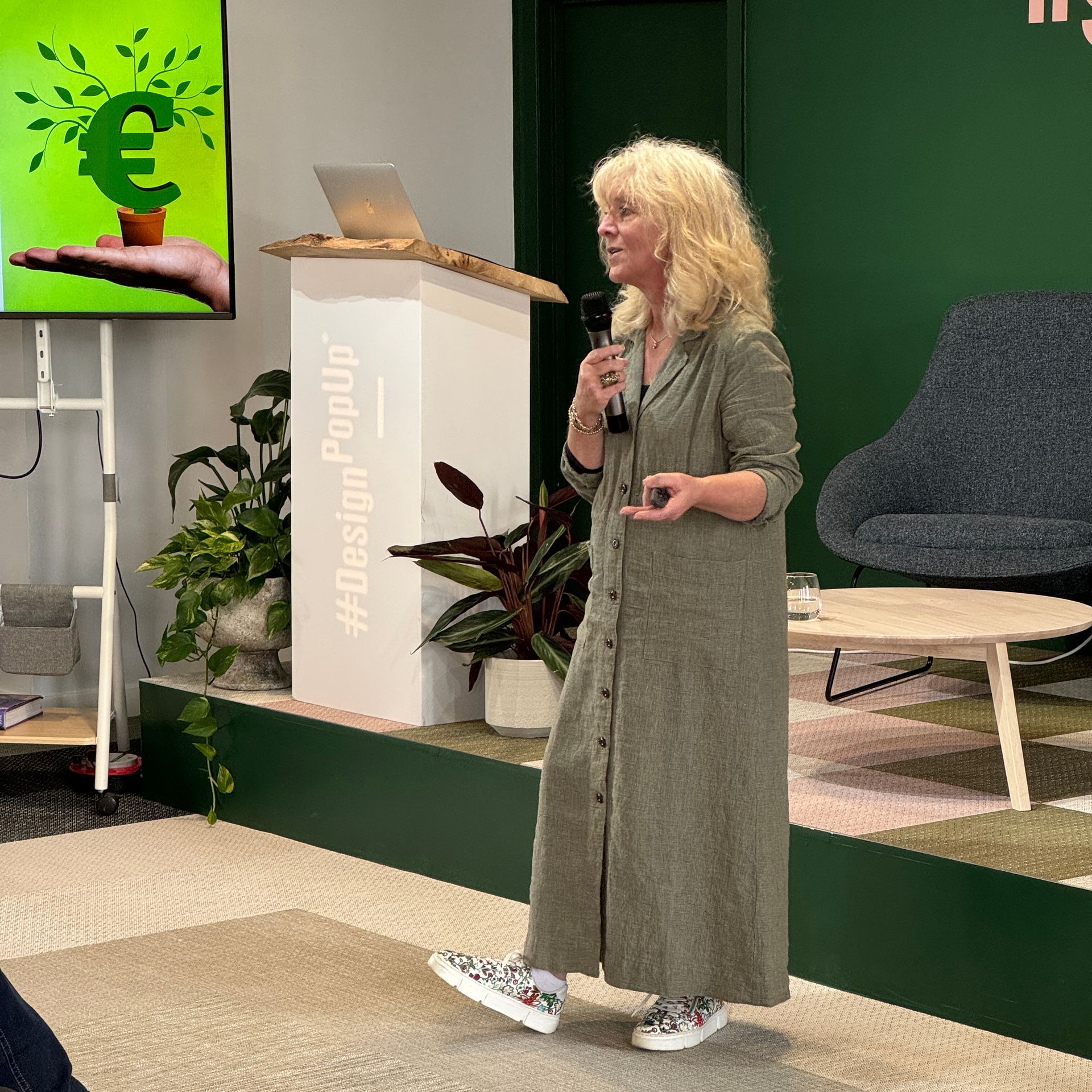 Woman giving a presentation at a design event; holding a microphone, wearing a long dress, standing by a podium.