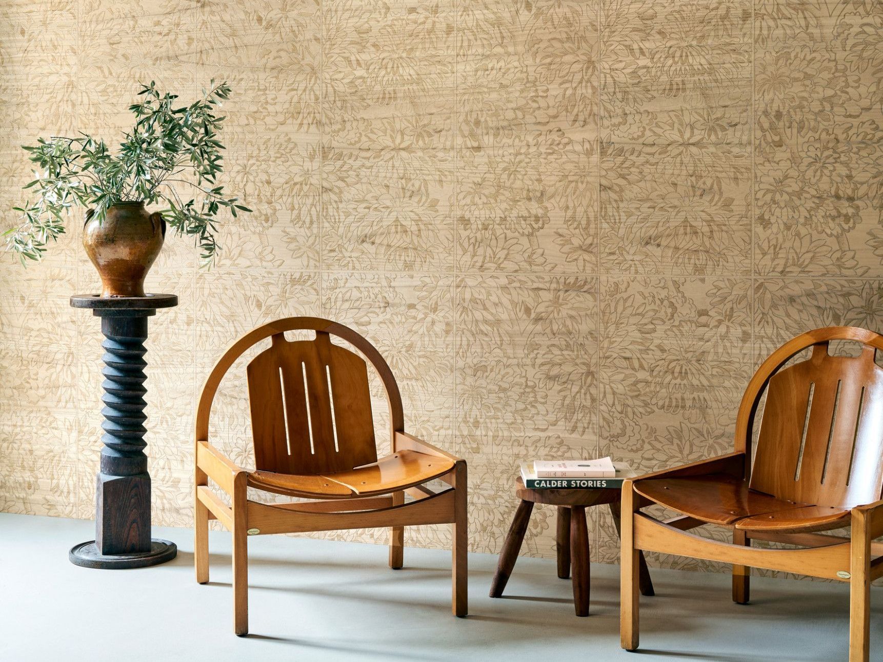 Two wooden chairs and a small stool in a room with textured walls and a plant on a pedestal.