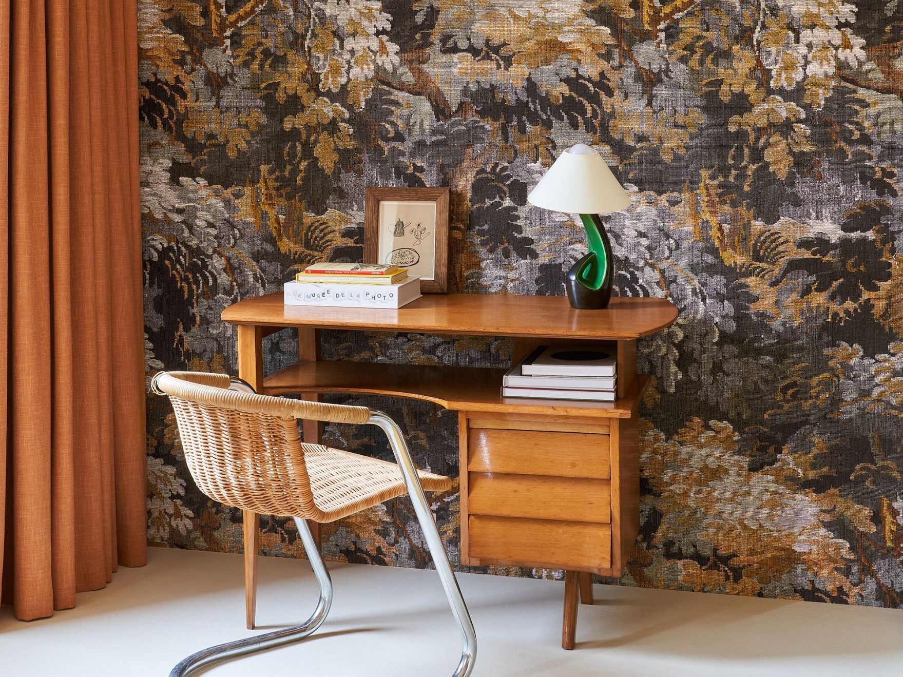 Mid-century desk with chair, lamp, and books against a forest-patterned wallpaper and orange curtain.