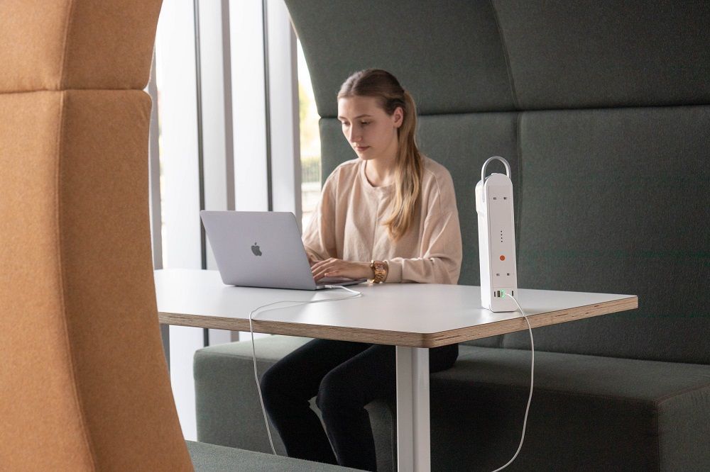 Woman typing on laptop at a desk in a booth; white power strip on table.