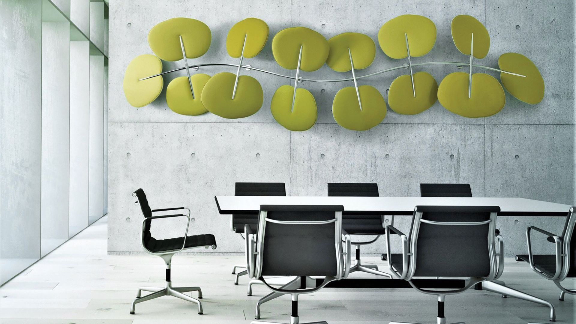 there is a modern table in a concrete office with natural light coming in from the large windows. at the table are seven black desk chairs. on the wall, there is a large leaf-like scultpure; it is green and large. 