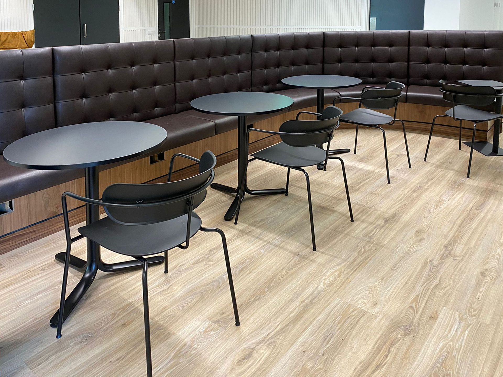 Upholstered bench seating in a curved line with small circular dining tables placed along the seating. On the other side of the tables are black metallic chairs.