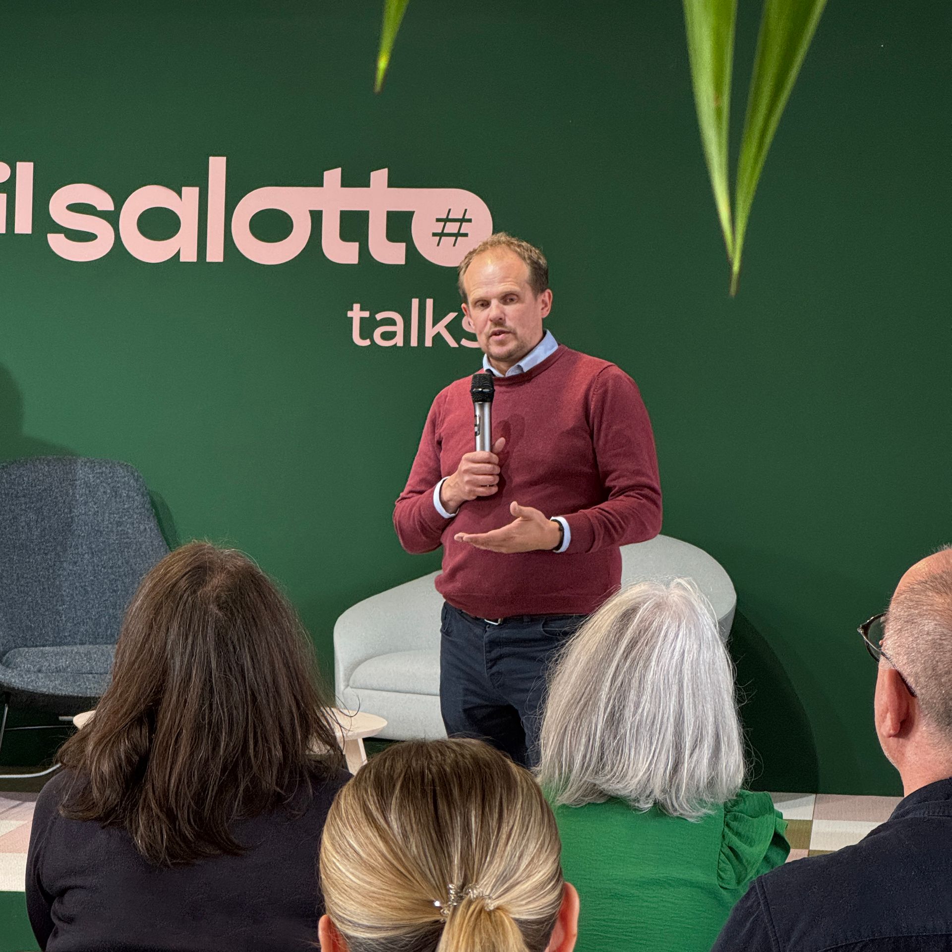 Man speaking into a microphone on stage, audience seated. Green backdrop with logo.