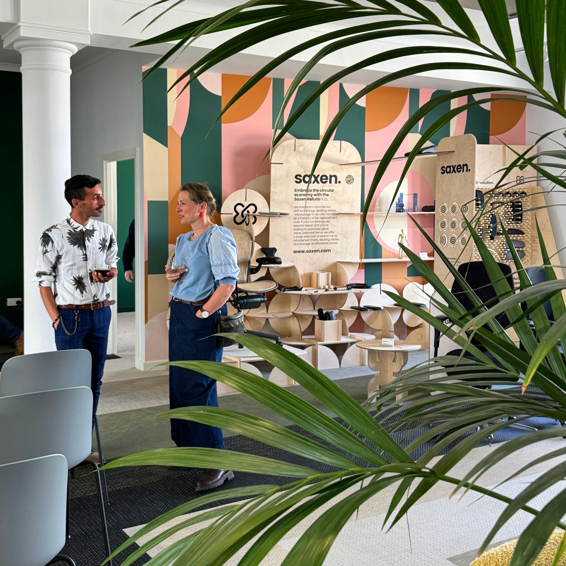 Two people converse near a Saxen commercial furniture display, with colourful wall art and potted plants in an airy room.