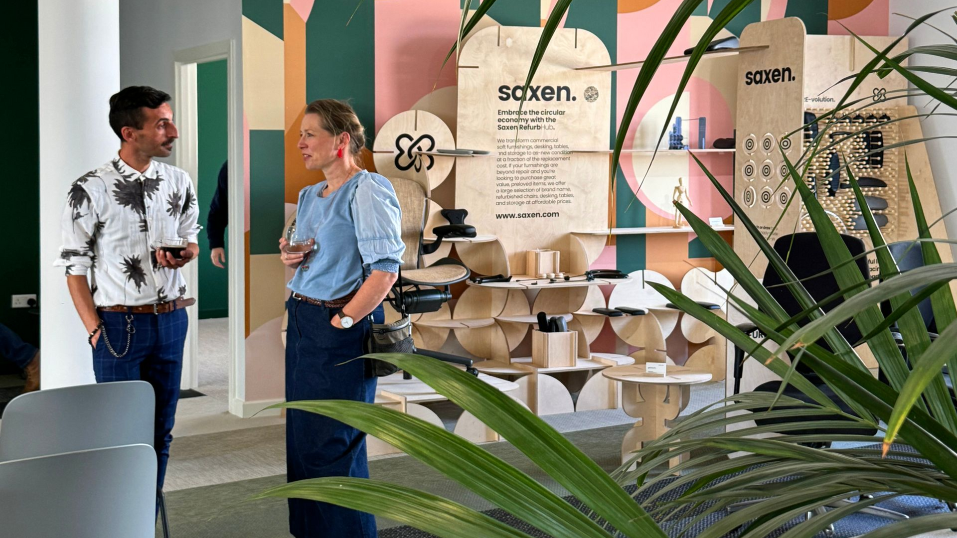 Two people conversing in a colorful room with display of wooden objects and plants.