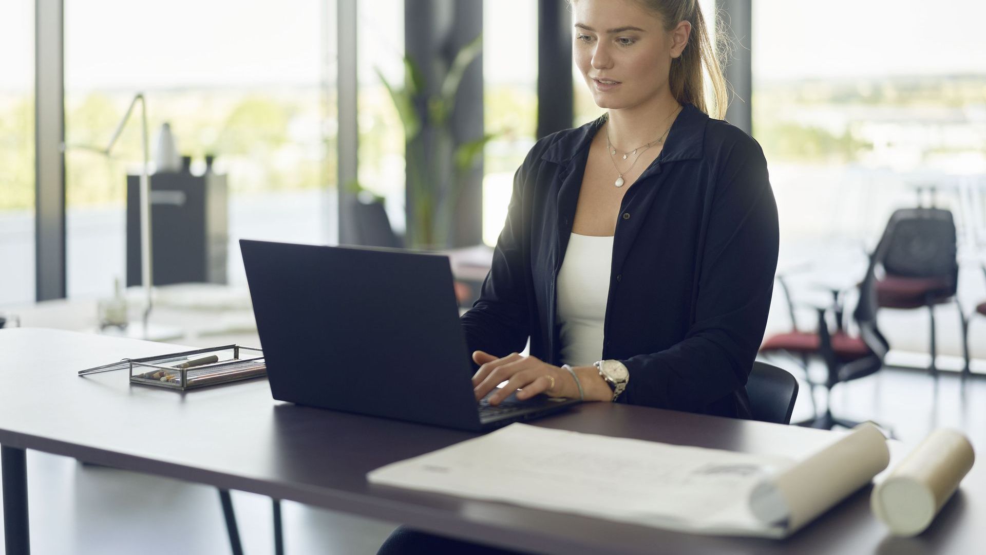 woman sitting at a desk, typing on her laptop. she is wearing a white top underneath a black button up shirt. t