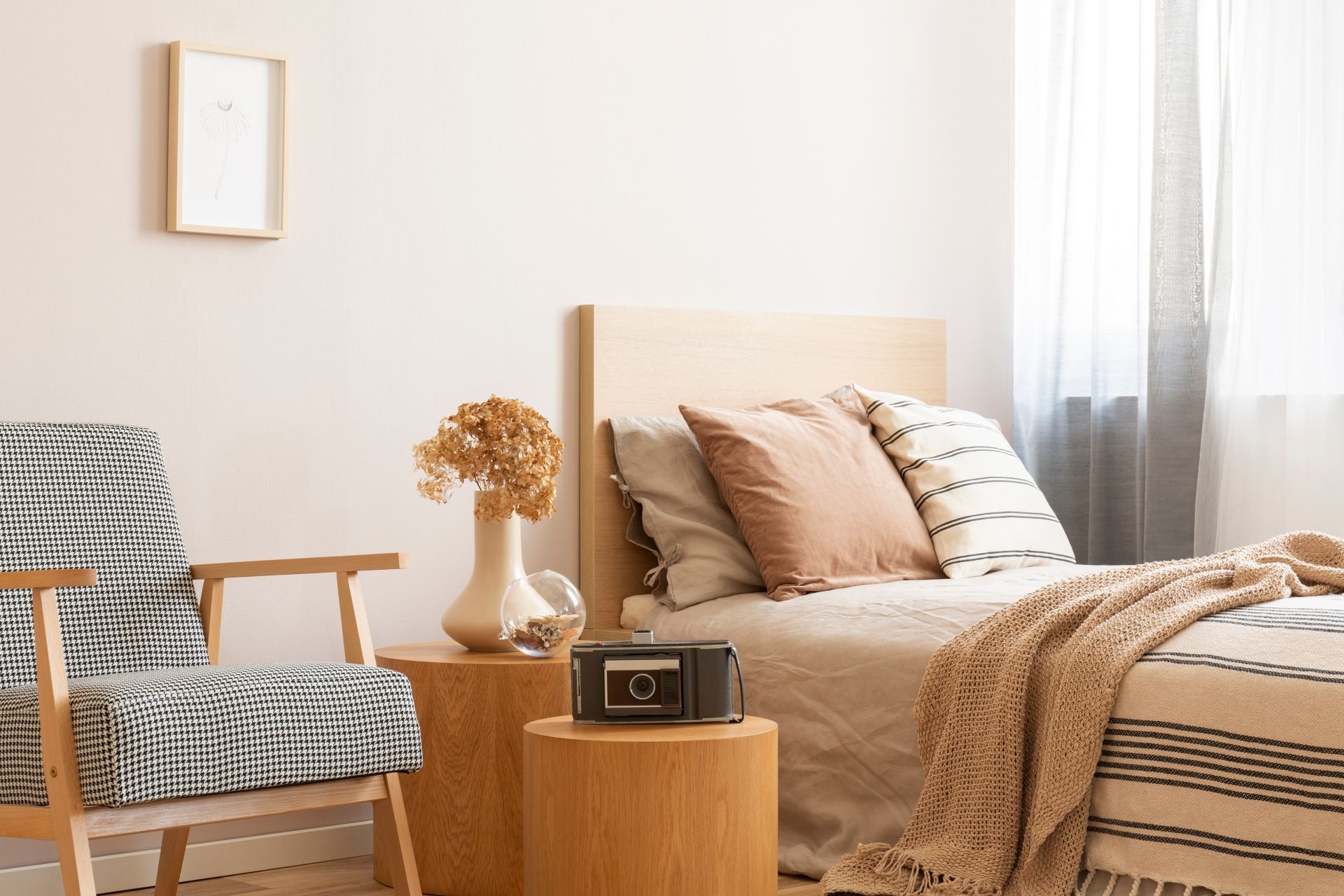A bedroom corner featuring a patterned armchair, a wooden side table with a vase and camera, and a bed with neutral linens.