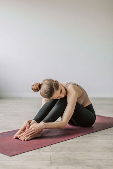 Persona estirándose en una postura de yoga sentada sobre una esterilla color granate en interiores