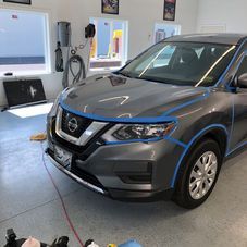 A grey Nissan Rogue SUV in a garage, with blue masking tape applied to the trim in preparation for detailing.