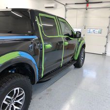 A black pickup truck parked in a garage with green and blue masking tape applied to the trim and windows for detailing.