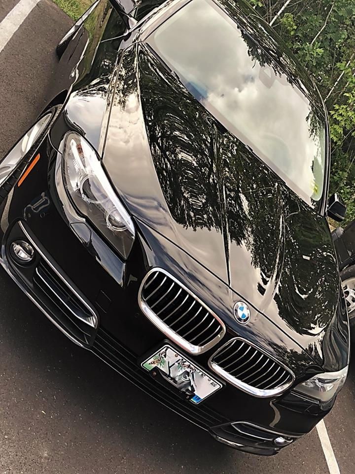 A black BMW sedan parked at an angle on an asphalt surface with trees reflected on the hood.