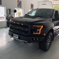 A black Ford F-150 Raptor truck with amber grille and headlight accents parked inside a brightly lit, clean garage.