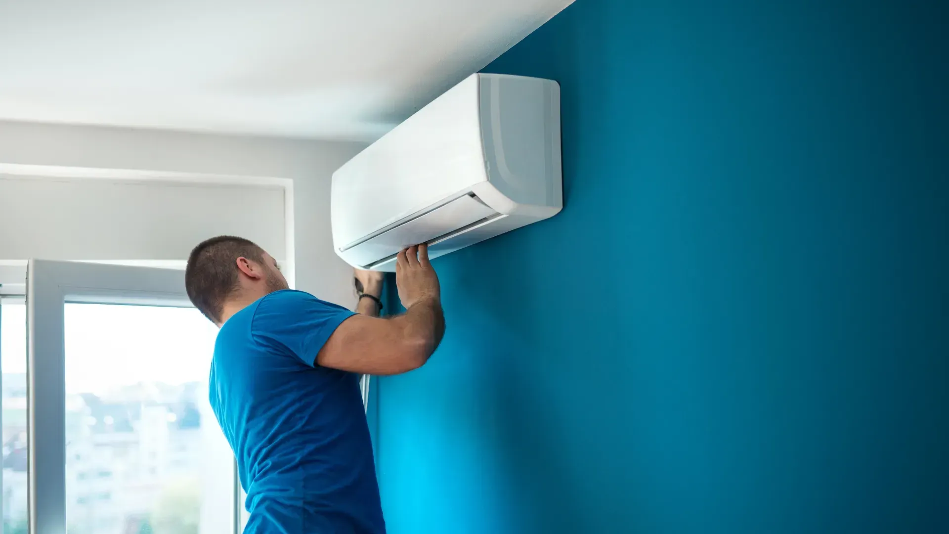 A man is installing an air conditioner on a blue wall.