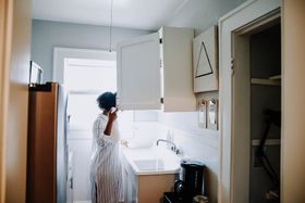 Person in white dress standing at a kitchen sink in a bright, open cabinet kitchen