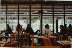 People seated at tables inside a warmly lit café with large glass windows.