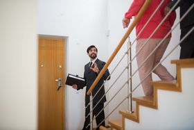 Man in a dark suit carries a box near a wooden staircase as another person in a red shirt descends.