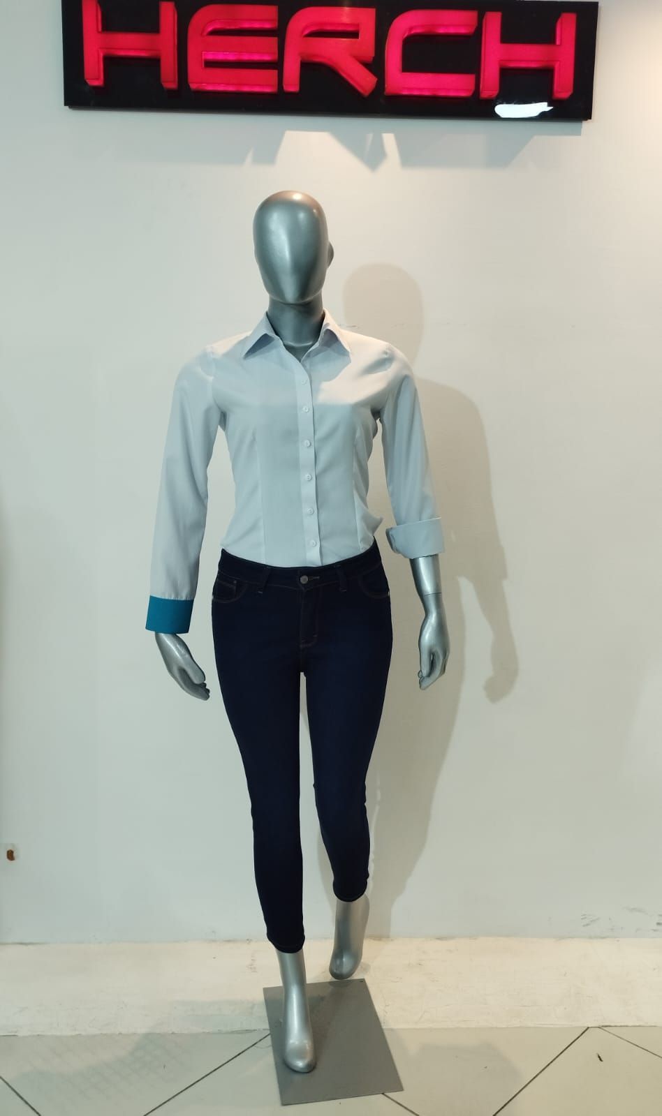 Maniquí con blusa azul claro abotonada, jeans oscuros y zapatos plateados; debajo de un cartel 