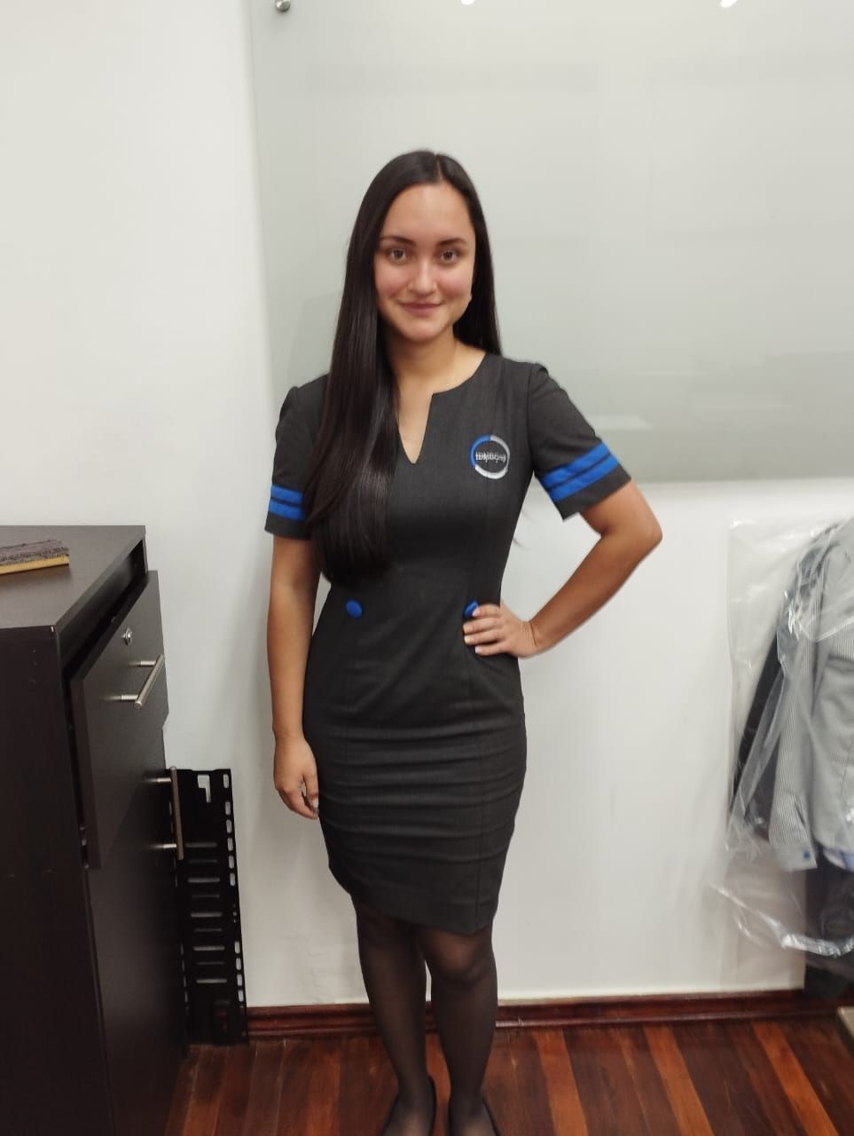 Mujer en uniforme negro, de pie, con la mano en la cadera, en un entorno de oficina.